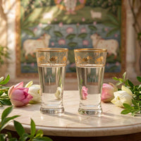 Two clear glasses with gold accents on a table with flowers and a decorative background.
