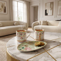 Two white ceramic mugs with pink floral and gold leaf patterns sit on a marble coffee table in a bright, elegant living room. A matching decorative plate with cookies rests on the table, and steam rises from the mugs.
