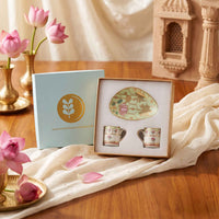 A boxed tea or coffee set displayed on a wooden table with draped cream fabric and pink lotus flowers. The set includes a light blue gift box with a gold leaf logo, an oval plate with floral artwork, and two small cups with floral patterns.
