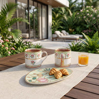 Two decorative ceramic mugs with floral and gold patterns sit on a wooden table outdoors, alongside a glass of orange juice and a plate of croissants on a beige runner. A modern patio with lounge chairs and lush greenery is visible in the background.