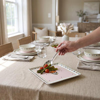 A hand uses a spoon to serve food from a square pink ceramic serving platter with a decorative border, set on a linen tablecloth in a dining room.