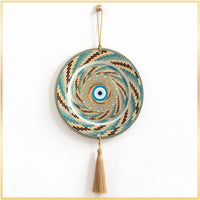 A circular woven wall hanging with a spiral pattern in teal, beige, brown, and white, featuring a central blue and white evil eye motif. A tan tassel hangs from the bottom center, and the piece is suspended by a rope loop at the top against a white background.