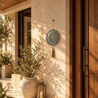 A circular blue and white patterned wall hanging with a tassel is displayed on a beige stucco wall next to a wooden door, with potted plants and warm sunlight casting shadows in the background.