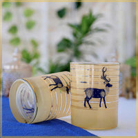 Two clear glass tumblers with a frosted pale yellow tint, decorated with horizontal gold stripes and a dark navy blue deer motif, displayed on a blue cloth with a blurred background.