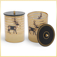 Two cylindrical decorative containers with lids, shown on a white background with a thin gold border. The containers are a warm beige/tan color with horizontal metallic gold stripes and a black deer motif.