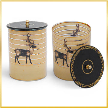 Two cylindrical decorative containers with lids, shown on a white background with a thin gold border. The containers are a warm beige/tan color with horizontal metallic gold stripes and a black deer motif.