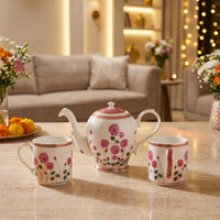 A porcelain tea set with a teapot and two matching cups decorated with pink floral patterns and gold trim, displayed on a marble countertop in a warm, modern living room setting.