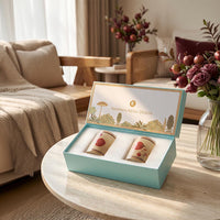 An open light blue gift box on a round table in a softly lit living room, containing two tumbler glasses with pomegrante illustrations. 