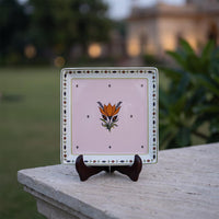 A square decorative ceramic plate with a light pink center, an orange flower motif, and a patterned border, displayed on a small dark wooden stand on a stone ledge with a blurred garden background.