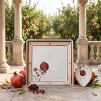 A decorative square ceramic plate with a pomegranate illustration, displayed alongside a matching heart-shaped bowl on a stone patio in front of an outdoor garden with columns.