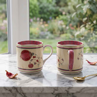Two ceramic mugs with cream backgrounds and red pomegranate illustrations, set on a white marble windowsill with a blurred garden view in the background.