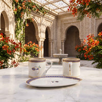 A white porcelain tea or coffee set with a matching saucer, featuring blue and gold patterned borders and a peacock motif, displayed on a white marble table in an ornate courtyard setting.