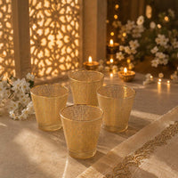 Four gold-toned textured glass candle holders arranged on a draped tablecloth with warm candlelight, floral decor, and a patterned screen in the background.