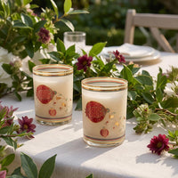 Two frosted tumbler glasses with gold trim and red pomegranate illustrations, styled on a white tablecloth outdoors with green leaves and flowers.