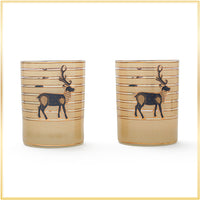 Two matching glasses tumblers with a warm beige gradient base, horizontal gold stripes, and a dark blue reindeer illustration on the front, set against a white background with a thin gold border.