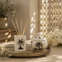 A stoneware reed diffuser and matching lidded jar with black palm tree illustrations, displayed on a decorative tray in a warm, sunlit room with flowers and a lattice screen in the background.