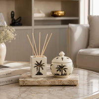 A decorative ceramic reed diffuser and matching lidded jar with black palm tree illustrations, displayed on a marble tray on a round table in a softly lit living room.