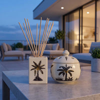 A reed diffuser and matching decorative jar with black palm tree illustrations, displayed on a stone patio table at dusk with a modern outdoor lounge area in the background.