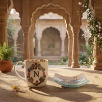 A ceramic mug and matching small plate set in a sunlit courtyard with stone arches. The mug is cream-colored with a floral wreath design and a large letter N, while the plate has a pale aqua rim and matching monogram details.
