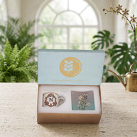 A personalized ceramic mug and matching coaster gift set presented in a kraft paper box with a light blue lid. The scene is set on a stone tabletop with a blurred background of a bright window, green plants, and a brass watering can.