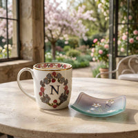 A white ceramic mug with a floral monogram Alphabet sits on a stone table beside a small matching glass dish, set against a soft-focus garden background.