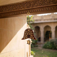 A decorative wooden hanging bell ornament with gold floral engravings and a small brass bell, suspended from a carved wooden beam in a sunlit courtyard.