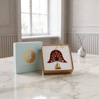 A decorative hanging ornament displayed in an open gift box on a marble tabletop, with a matching closed box standing beside it. The ornament has a dark red bell-shaped body with gold floral detailing and a small gold hanging loop and bell.